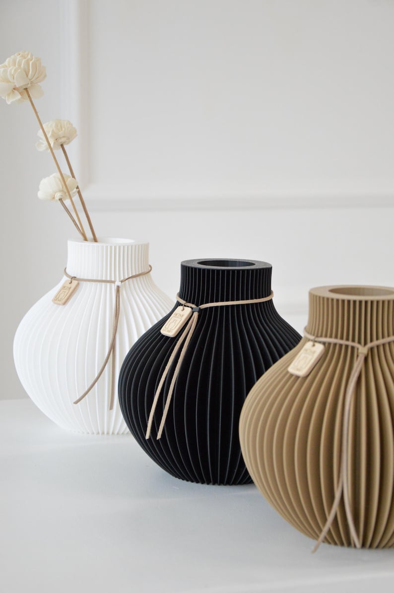 May include: Three decorative vases in white, black, and tan, each with a unique ribbed design. The white vase holds dried flowers. Each vase has a tag and is tied with a thin, light-colored cord. The vases are on a white surface.