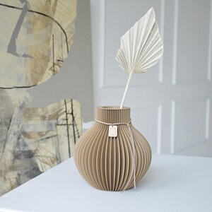 May include: A beige, ribbed vase with a dried palm leaf. The vase has a round shape and is tied with a pink ribbon and a small wooden tag. The vase is on a white surface.