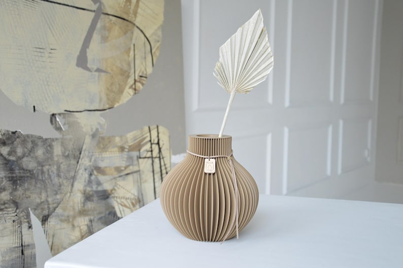 May include: A beige, ribbed vase with a dried palm leaf. The vase has a round shape and is tied with a pink ribbon and a small wooden tag. The vase is on a white surface.