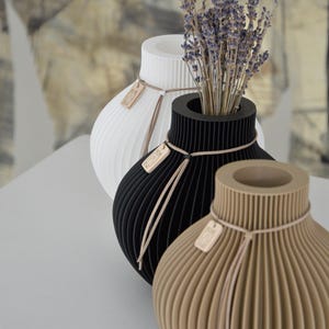 May include: Three decorative vases in white, black, and tan, each with a unique ribbed design. The black vase holds dried lavender. Each vase has a small wooden tag tied with twine. The vases are approximately 8 inches tall.