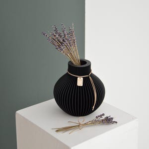 May include: A black, spherical vase with vertical grooves, holding dried lavender. A small, rectangular tag hangs from a string tied around the vase. A small bundle of lavender rests on the white surface. The background is split between white and a muted green.