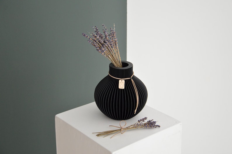 May include: A black, spherical vase with vertical grooves, holding dried lavender. A small, rectangular tag hangs from a string tied around the vase. A small bundle of lavender rests on the white surface. The background is split between white and a muted green.