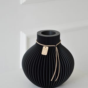 May include: Black decorative vase with a unique, ribbed design. The vase has a rounded body and a cylindrical neck. A small wooden tag with text hangs from a tan string tied around the neck. The vase is displayed against a white background.