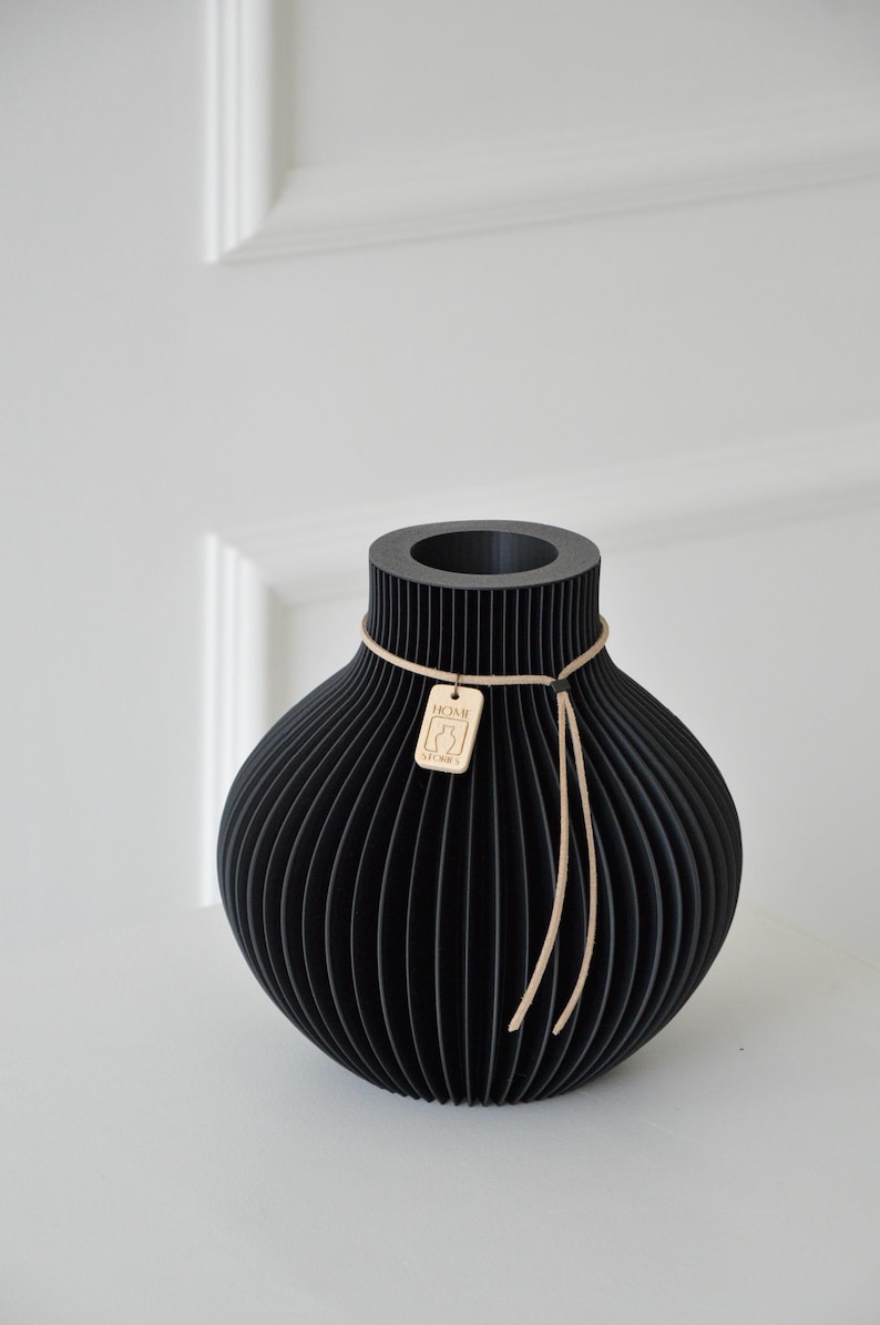 May include: Black decorative vase with a unique, ribbed design. The vase has a rounded body and a cylindrical neck. A small wooden tag with text hangs from a tan string tied around the neck. The vase is displayed against a white background.