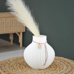 May include: A white, ribbed ceramic vase with a decorative cord and wooden charm, holding a tall, feathery, cream-colored plume. The vase sits on a woven, circular mat, with a dark green background.