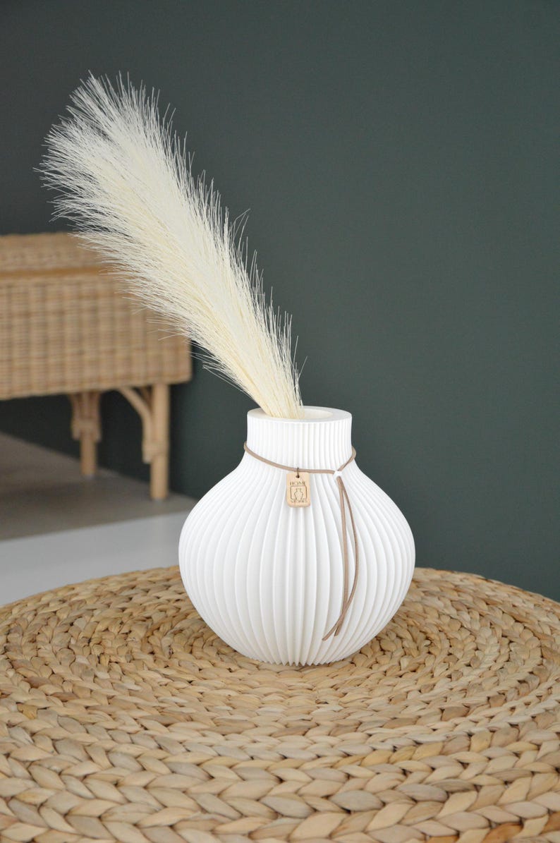 May include: A white, ribbed ceramic vase with a decorative cord and wooden charm, holding a tall, feathery, cream-colored plume. The vase sits on a woven, circular mat, with a dark green background.