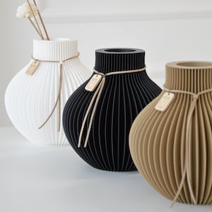 May include: Three decorative vases in white, black, and tan. Each vase has a unique ribbed design and is adorned with a small wooden tag tied with a string. The white vase holds dried flowers. A modern home decor item.