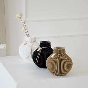 May include: Three decorative vases in white, black, and tan, each with a unique ribbed design. The white vase holds a few white flowers. Each vase has a small tag attached with twine. The vases are displayed on a white surface.