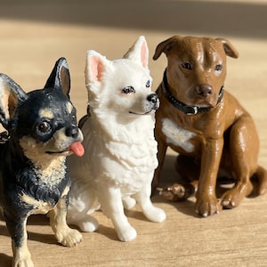 May include: Three dog figurines: a black and tan Chihuahua with its tongue out, a white fluffy dog, and a brown dog with a black collar. The dogs are sitting on a light brown surface.