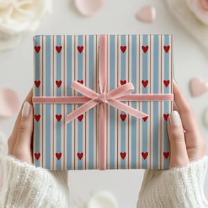 May include: A wrapped gift box with a blue and white striped pattern and red hearts. The box is tied with a pink velvet ribbon and bow. The gift is held by a person wearing a white sweater.