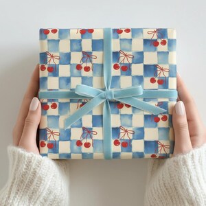 May include: A wrapped gift with a blue and white checkered pattern featuring red cherries. The gift is tied with a light blue velvet ribbon and bow. The gift is held by a person wearing a white sweater.