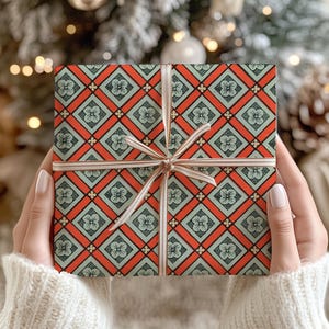 May include: A square gift box wrapped in paper featuring a repeating geometric pattern of orange diamonds and light green squares with small floral motifs, tied with a thin, light-coloured ribbon centred in a bow across the front surface.