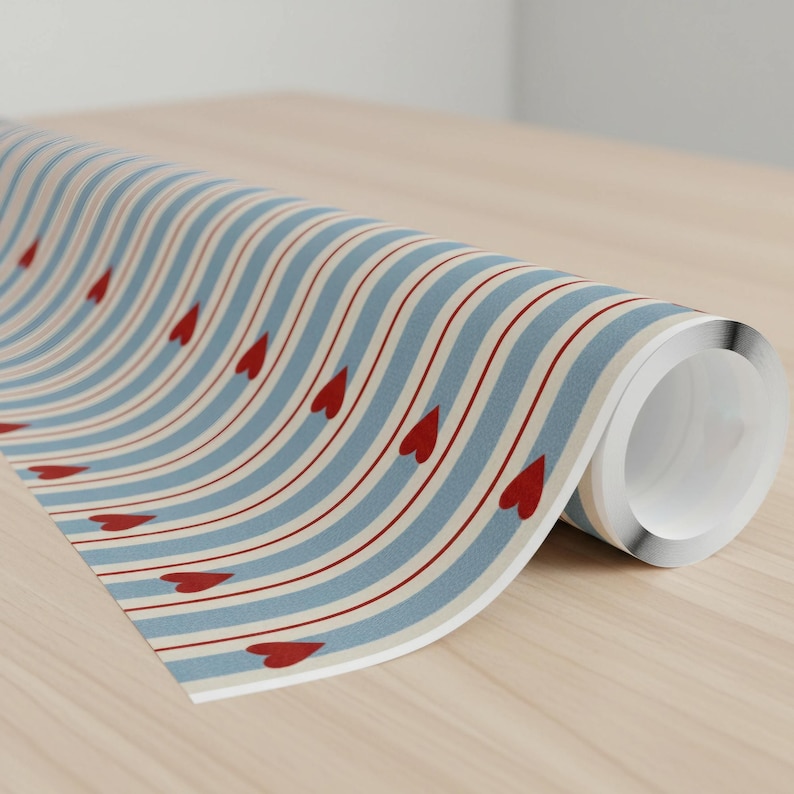 May include: Rolled-up patterned paper featuring a repeating design of red hearts and alternating light blue and off-white wavy stripes. The paper is on a light wood surface.