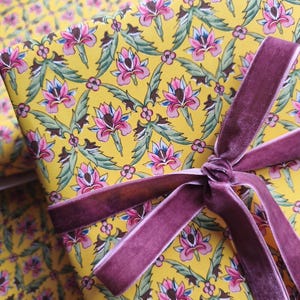 May include: A yellow gift wrap paper featuring a repeating pattern of pink and blue floral motifs with green leaves, tied with a dark mauve velvet ribbon. The ribbon is knotted in a bow across the surface of the patterned paper.