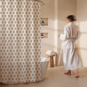 May include: A beige shower curtain with a repeating floral pattern in muted red and green. The curtain hangs from a curved metal rod. The bathroom has a white tub, wooden stool, and a person wearing a white robe.