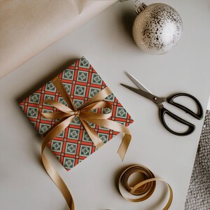 May include: A square gift box wrapped in paper with a repeating grid pattern of red lines and light blue floral squares, tied with a gold satin ribbon. Nearby are black metal scissors, a silver glittery ornament, and a spool of gold ribbon.