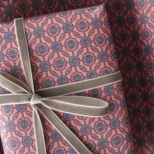 May include: A rectangular gift wrapped in pink paper featuring a repeating dark blue floral and geometric lattice pattern. The package is tied with a smooth, light gray velvet ribbon that is knotted into a bow on the front surface.