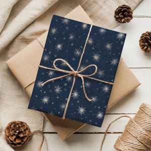 May include: A wrapped gift with navy blue paper featuring a white starburst pattern. The package is tied with twine and rests on a brown paper-wrapped box. Pinecones and a spool of twine are also visible.