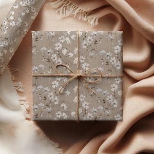May include: A square gift box wrapped in floral patterned paper with white flowers on a tan background. The box is tied with twine and a bow. A roll of matching wrapping paper is visible in the upper left corner.