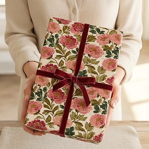May include: A wrapped gift with a floral pattern of pink peonies and green leaves on an off-white background. The gift is tied with a burgundy velvet ribbon and bow, creating a classic and elegant presentation.