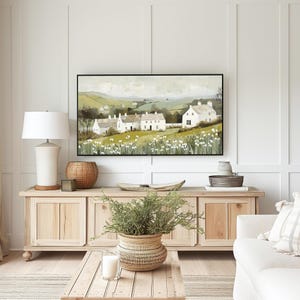 May include: A framed painting depicting a countryside scene with white houses and green hills. The painting is hung above a wooden cabinet with a plant in a woven basket.