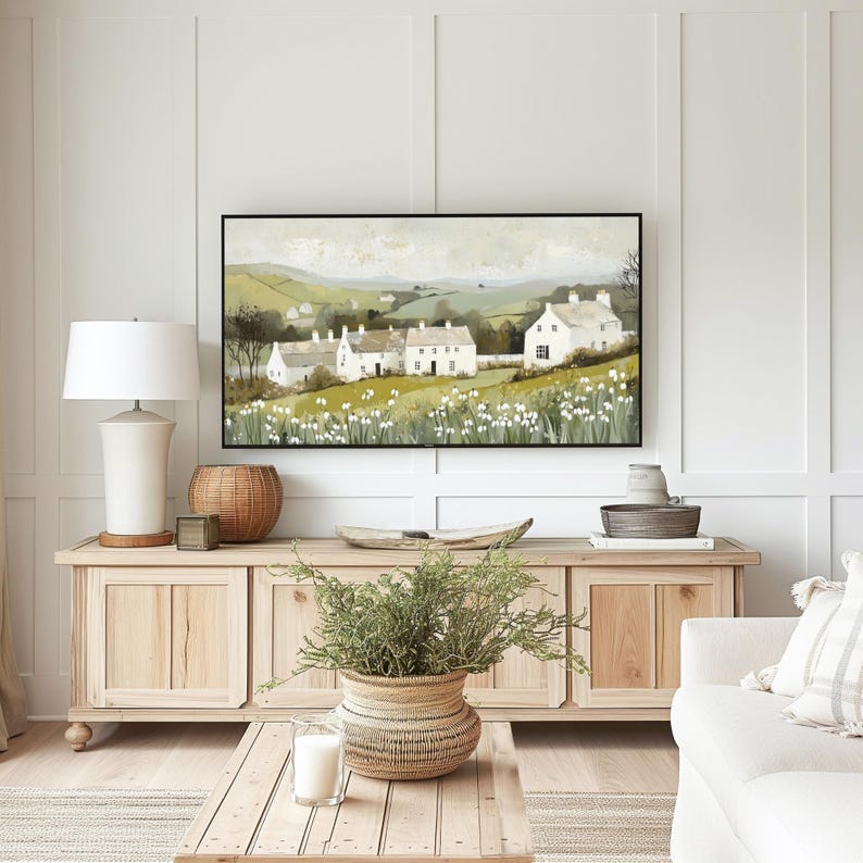 May include: A framed painting depicting a countryside scene with white houses and green hills. The painting is hung above a wooden cabinet with a plant in a woven basket.