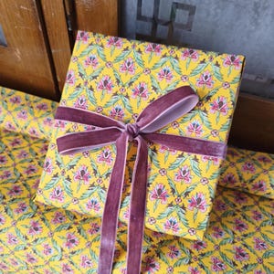 May include: A square gift box wrapped in bright yellow paper featuring a repeating floral pattern of pink and white blossoms with green leaves. The box is tied with a wide, dark mauve velvet ribbon that is knotted in a bow at the center.