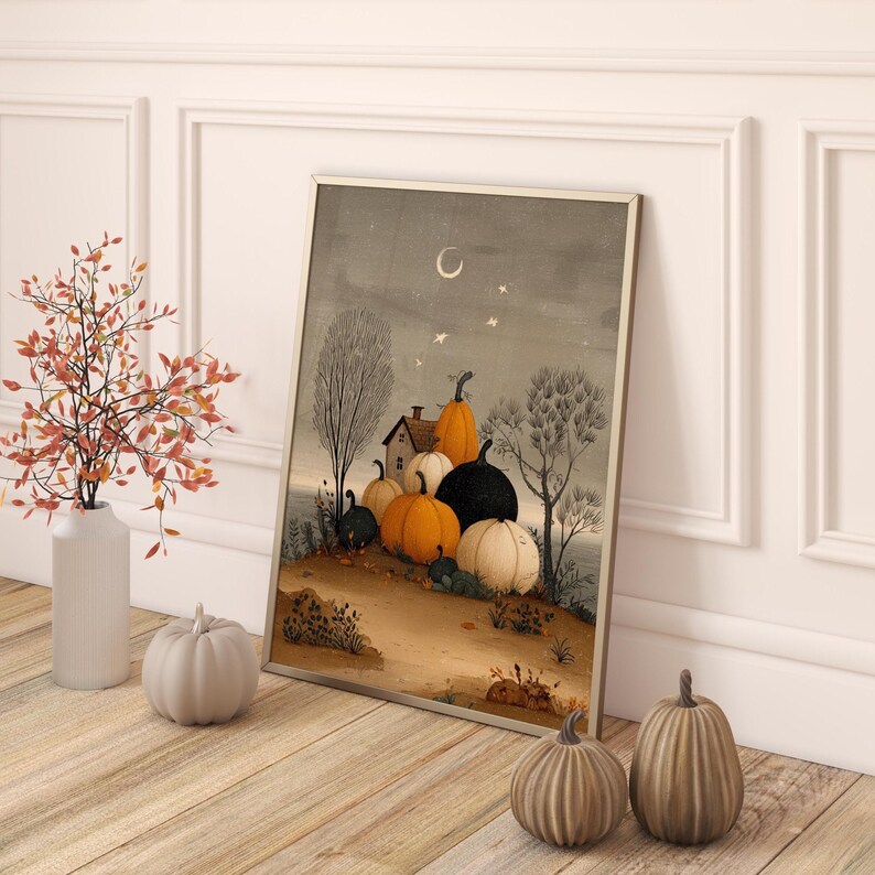 May include: A framed art print depicting a harvest scene with pumpkins, a house, and trees under a night sky with a crescent moon and stars. Decorative pumpkins in various sizes and colours are in front of the print. A vase with autumn foliage is on the left.