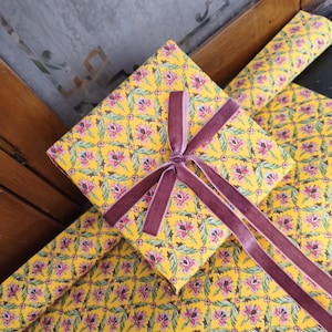 May include: A square gift box wrapped in bright yellow paper featuring a repeating floral pattern of pink lotus-like flowers and green leaves. The box is tied with a dark mauve velvet ribbon that is knotted in the center with four long trailing ends.