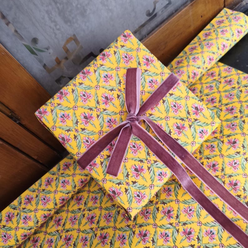 May include: A square gift box wrapped in bright yellow paper featuring a repeating floral pattern of pink lotus-like flowers and green leaves. The box is tied with a dark mauve velvet ribbon that is knotted in the center with four long trailing ends.