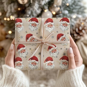 May include: A wrapped gift box featuring a repeating pattern of Santa Claus faces in red hats and white beards on a beige background. The box is tied with a brown ribbon and bow. The gift is held by a person wearing a white sweater.