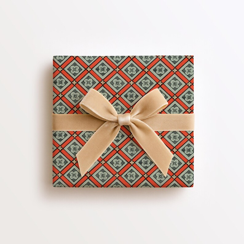 May include: A square gift box wrapped in paper featuring a repeating diamond pattern in orange and muted green, with each diamond containing a small floral motif. A wide, smooth, beige velvet ribbon is tied in a bow across the centre.