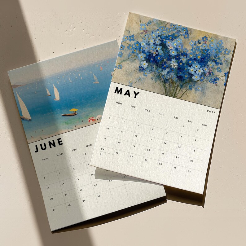 May include: Two wall calendar pages for May and June 2027. The May page features a blue floral bouquet painting, while the June page displays a coastal scene with sailboats on the water. Both pages have black text headers and a grid layout.