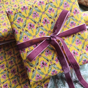 May include: A rectangular gift box wrapped in bright yellow paper featuring a repeating floral pattern of pink and blue blossoms with green leaves. The box is tied with a dark mauve velvet ribbon tied into a bow on the top surface.