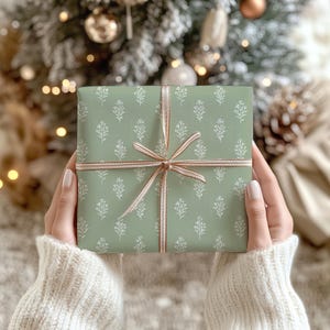 May include: A wrapped gift with sage green paper featuring a white floral pattern. The gift is tied with a light brown and white striped ribbon. The background includes a Christmas tree with soft, blurred lights.