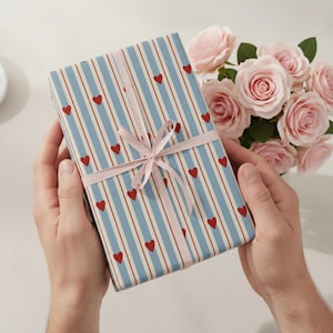 May include: A wrapped gift with blue and white striped paper, red heart accents, and a pink ribbon. A bouquet of pink roses is in the background. The gift is held in both hands.