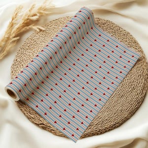 May include: A roll of patterned paper featuring a repeating design of red hearts and red stripes on a light blue background. The paper is rolled up and placed on a woven, circular mat.