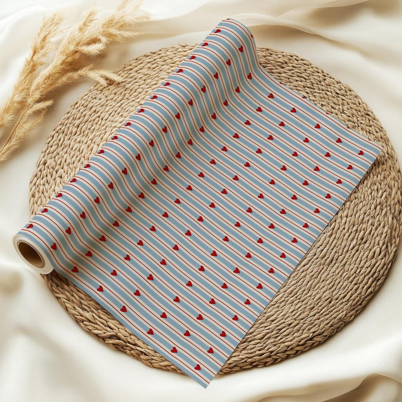 May include: A roll of patterned paper featuring a repeating design of red hearts and red stripes on a light blue background. The paper is rolled up and placed on a woven, circular mat.