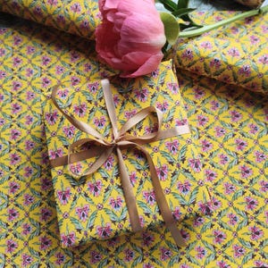 May include: A rectangular gift box wrapped in bright yellow paper featuring a repeating floral pattern of pink blossoms and green leaves, tied with a smooth tan ribbon bow, accompanied by a single large pink peony flower resting on top.
