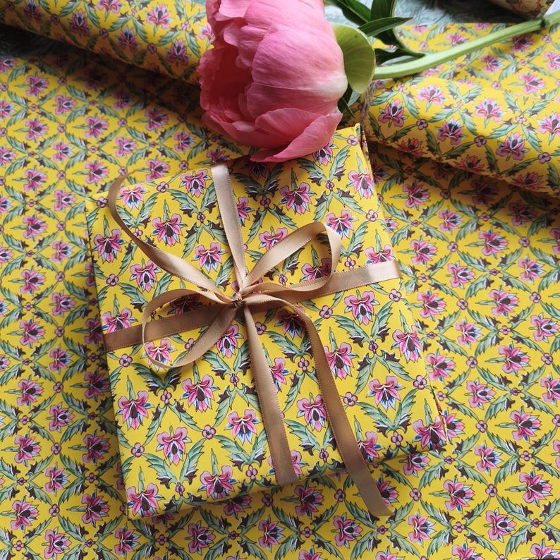May include: A rectangular gift box wrapped in bright yellow paper featuring a repeating floral pattern of pink blossoms and green leaves, tied with a smooth tan ribbon bow, accompanied by a single large pink peony flower resting on top.