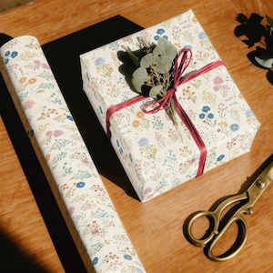 May include: A wrapped gift box and a roll of floral wrapping paper. The paper has a cream background with small blue, yellow, and pink flowers. The gift is tied with a red ribbon and decorated with greenery. Gold scissors are on the wooden surface.