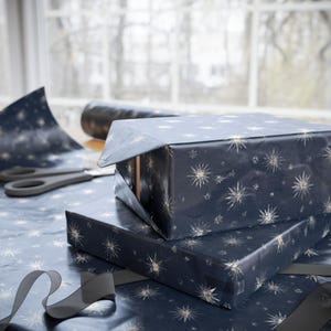 May include: Two wrapped gifts are stacked on a table, covered in navy blue paper with a gold starburst pattern. A pair of scissors and a roll of matching wrapping paper are nearby. A dark gray ribbon is draped across the table.