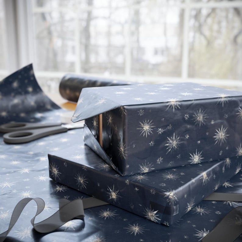 May include: Two wrapped gifts are stacked on a table, covered in navy blue paper with a gold starburst pattern. A pair of scissors and a roll of matching wrapping paper are nearby. A dark gray ribbon is draped across the table.