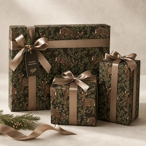May include: Three rectangular gift boxes wrapped in dark paper featuring a repeating pattern of spotted fawns and green foliage. Each box is tied with a smooth, light brown satin ribbon. A dark tag with the text Season's Greetings is attached.
