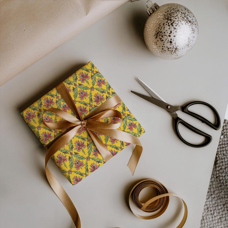 May include: A square gift box wrapped in yellow paper with a repeating floral pattern of pink flowers and green leaves, tied with a gold satin ribbon bow. Nearby are black metal scissors, a silver glittery ornament, and a spool of gold ribbon.