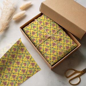 May include: A rectangular cardboard box containing an item wrapped in yellow paper with a repeating floral pattern of red flowers and green leaves, tied with thin brown twine. A matching sheet of yellow floral wrapping paper lies nearby.