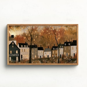 May include: A rectangular framed wall art print depicting a row of houses in a wooded setting during autumn. The scene features dark and light-coloured buildings with glowing windows, surrounded by trees with orange foliage and string lights.