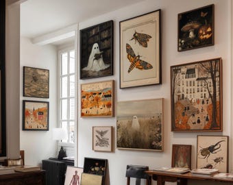 May include: A gallery wall featuring framed art prints with various themes. The collection includes illustrations of a ghost reading, moths, a mushroom and pumpkin, and a cityscape. The frames are in various sizes and styles, creating a curated display.