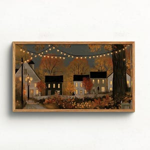 May include: A framed horizontal wall art print depicting a neighborhood scene at dusk with autumn foliage, several houses with glowing windows, a large tree trunk on the right, and a string of warm white fairy lights hanging across the dark sky.