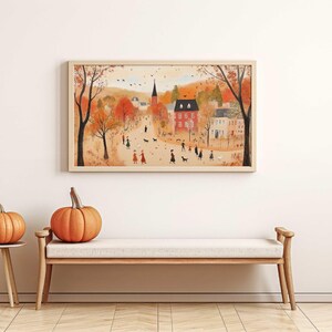 K&ouml;nnte beinhalten: Eine gerahmte Illustration einer kleinen Stadt mit einem Kirchturm, roten Backsteingeb&auml;uden und Menschen, die auf einem von B&auml;umen ges&auml;umten Weg spazieren gehen. Die B&auml;ume haben orange, gelbe und rote Bl&auml;tter, was auf den Herbst hindeutet.